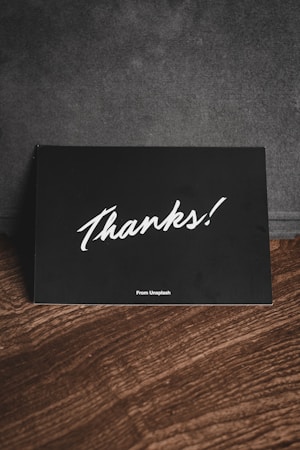 A black card with white handwriting displaying the word 'Thanks!' is placed on a wooden surface. There is a subtle text 'From Unsplash' at the bottom, and the background appears to be a dark fabric material.