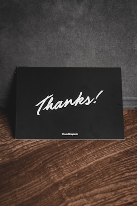 A black card with white handwriting displaying the word 'Thanks!' is placed on a wooden surface. There is a subtle text 'From Unsplash' at the bottom, and the background appears to be a dark fabric material.