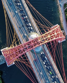 An aerial view of a large suspension bridge with red cables spanning over a river. Multiple lanes of traffic are visible, including a variety of cars traveling in both directions. The bridge incorporates a geometric central hub, possibly observatory or lookout, with a glass dome. The structure extends over dark blue water, with a pathway visible on the riverbank.