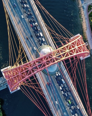 An aerial view of a large suspension bridge with red cables spanning over a river. Multiple lanes of traffic are visible, including a variety of cars traveling in both directions. The bridge incorporates a geometric central hub, possibly observatory or lookout, with a glass dome. The structure extends over dark blue water, with a pathway visible on the riverbank.