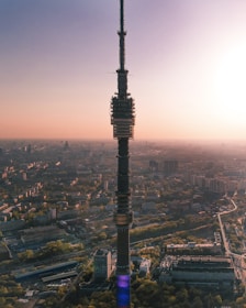 A tall broadcasting or telecommunications tower stands prominently in the center of an expansive urban landscape during sunset. The tower is surrounded by a sprawling city with buildings, residential areas, and tree-lined streets, all bathed in a warm, golden light. The horizon is slightly hazy, with the sun positioned just off to the side, creating a gradient in the sky from light gold to soft pink.