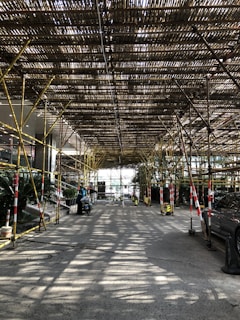 A structure made of bamboo scaffolding covers a roadway, allowing light to filter through, creating striped patterns on the ground. There are several vehicles parked along the sides, and a few pedestrians are visible. The scene is framed by trees and plants at the edges.