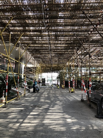 A structure made of bamboo scaffolding covers a roadway, allowing light to filter through, creating striped patterns on the ground. There are several vehicles parked along the sides, and a few pedestrians are visible. The scene is framed by trees and plants at the edges.