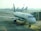 A row of airplanes is parked at an airport gate area, with a focus on the Alitalia aircraft situated closest to the camera. The airplane is connected to a passenger boarding bridge. Several other airplanes are visible in the background, lined up along the tarmac under the overcast sky.