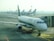 A row of airplanes is parked at an airport gate area, with a focus on the Alitalia aircraft situated closest to the camera. The airplane is connected to a passenger boarding bridge. Several other airplanes are visible in the background, lined up along the tarmac under the overcast sky.