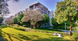 A modern building surrounded by lush green trees and a well-maintained lawn. Stone pathways and landscaped gardens enhance the visuals, while bright daylight highlights the serene atmosphere.