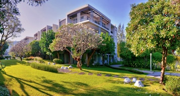 A modern building surrounded by lush green trees and a well-maintained lawn. Stone pathways and landscaped gardens enhance the visuals, while bright daylight highlights the serene atmosphere.