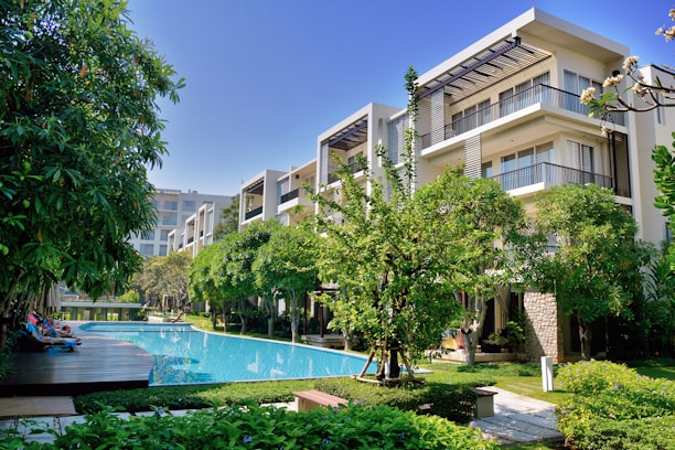 A modern apartment complex surrounded by lush greenery, featuring a clear blue swimming pool at the center. The buildings have a sleek design with large windows and balcony areas. People are leisurely lounging poolside on deck chairs, creating a relaxing and upscale atmosphere.
