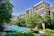 A modern apartment complex surrounded by lush greenery, featuring a clear blue swimming pool at the center. The buildings have a sleek design with large windows and balcony areas. People are leisurely lounging poolside on deck chairs, creating a relaxing and upscale atmosphere.