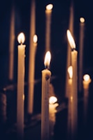Multiple lit candles with melting wax are arranged in a dark, dimly lit setting, casting a warm glow and soft shadows, creating a peaceful and tranquil atmosphere.