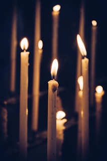 Multiple lit candles with melting wax are arranged in a dark, dimly lit setting, casting a warm glow and soft shadows, creating a peaceful and tranquil atmosphere.