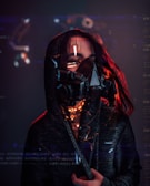 A person wearing a dark hooded cloak and a futuristic, black respirator mask with visible digital text and graphics superimposed around them. Red lighting highlights the edges and creates a dramatic contrast against the dark background.