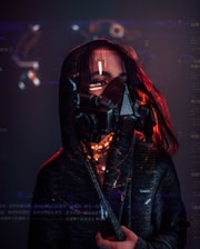 A person wearing a dark hooded cloak and a futuristic, black respirator mask with visible digital text and graphics superimposed around them. Red lighting highlights the edges and creates a dramatic contrast against the dark background.