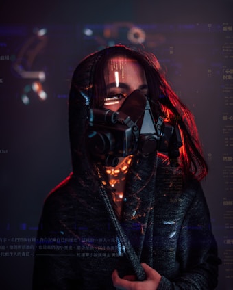 A person wearing a dark hooded cloak and a futuristic, black respirator mask with visible digital text and graphics superimposed around them. Red lighting highlights the edges and creates a dramatic contrast against the dark background.