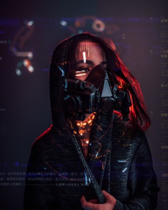 A person wearing a dark hooded cloak and a futuristic, black respirator mask with visible digital text and graphics superimposed around them. Red lighting highlights the edges and creates a dramatic contrast against the dark background.
