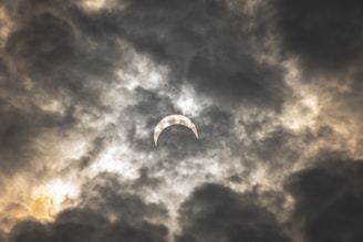 A solar eclipse partially obscures the sun, surrounded by dramatic, swirling clouds in varying shades of gray and white, with hints of warm yellow highlighting the edges.