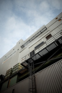 A tall industrial building with corrugated metal siding and various structural elements such as ladders and walkways. The perspective looks upward towards the sky, creating a dramatic angle. The building is marked with signs and logos.