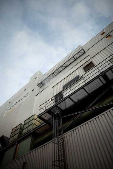 A tall industrial building with corrugated metal siding and various structural elements such as ladders and walkways. The perspective looks upward towards the sky, creating a dramatic angle. The building is marked with signs and logos.