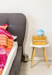 A modern bedroom setup featuring a gray upholstered bed with a vibrant pink and orange striped blanket. Next to the bed, a white and wooden bedside table holds a wooden box and a blue object with a digital screen.