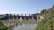 A large dam spans a river, with structures and gates controlling the water flow. The foreground features water and scattered rocks, while lush green vegetation lines the banks of the river. A clear blue sky forms the backdrop, and there is a large electricity pylon connected to power lines on the right, indicating industrial activity.