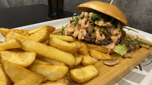 A gourmet hamburger is artistically presented on a wooden board alongside a generous portion of thick-cut fries. The burger is generously topped with greens, sautéed onions, mushrooms, and a creamy sauce, all sandwiched in a glossy bun.