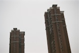 Two tall, brown residential towers with multiple windows stand against a pale, overcast sky. The buildings have simple, geometric architectural designs and feature some protruding ornamental structures.