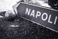 A weathered sign displaying the word 'Napoli' is resting on the ground, surrounded by grass and foliage. Beside it lies an old soccer ball, indicating a sense of nostalgia or abandoned activity.