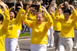 A group of people is standing outdoors performing coordinated arm movements. They are wearing matching yellow shirts and appear to be participating in a public event or demonstration. The individuals are arranged in rows, with some smiling, creating an inclusive and harmonious atmosphere.