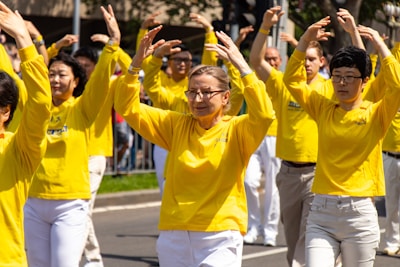 A group of people is standing outdoors performing coordinated arm movements. They are wearing matching yellow shirts and appear to be participating in a public event or demonstration. The individuals are arranged in rows, with some smiling, creating an inclusive and harmonious atmosphere.