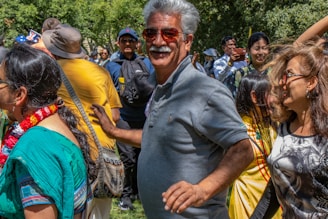 A group of people gathered outdoors, appearing cheerful and engaged in some activity. The scene includes a mix of individuals wearing colorful attire, and several are smiling or laughing. It's a sunny day, and some trees can be seen in the background.