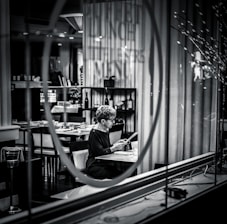A person sits alone at a table in a dimly lit restaurant, reading a menu. The setting is captured through a glass window with reflections, adding a sense of separation and introspection. Shelves with wine bottles and neatly arranged tables enhance the upscale atmosphere.