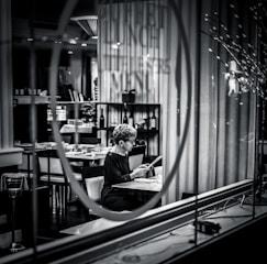 A person sits alone at a table in a dimly lit restaurant, reading a menu. The setting is captured through a glass window with reflections, adding a sense of separation and introspection. Shelves with wine bottles and neatly arranged tables enhance the upscale atmosphere.