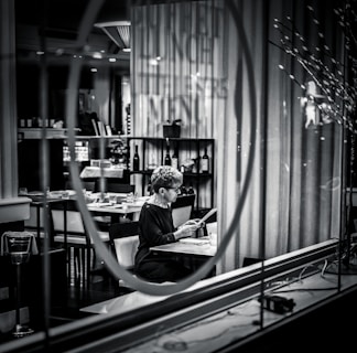 A person sits alone at a table in a dimly lit restaurant, reading a menu. The setting is captured through a glass window with reflections, adding a sense of separation and introspection. Shelves with wine bottles and neatly arranged tables enhance the upscale atmosphere.
