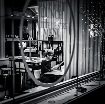 A person sits alone at a table in a dimly lit restaurant, reading a menu. The setting is captured through a glass window with reflections, adding a sense of separation and introspection. Shelves with wine bottles and neatly arranged tables enhance the upscale atmosphere.