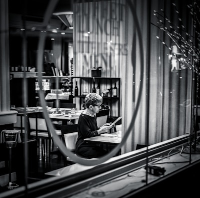 A person sits alone at a table in a dimly lit restaurant, reading a menu. The setting is captured through a glass window with reflections, adding a sense of separation and introspection. Shelves with wine bottles and neatly arranged tables enhance the upscale atmosphere.