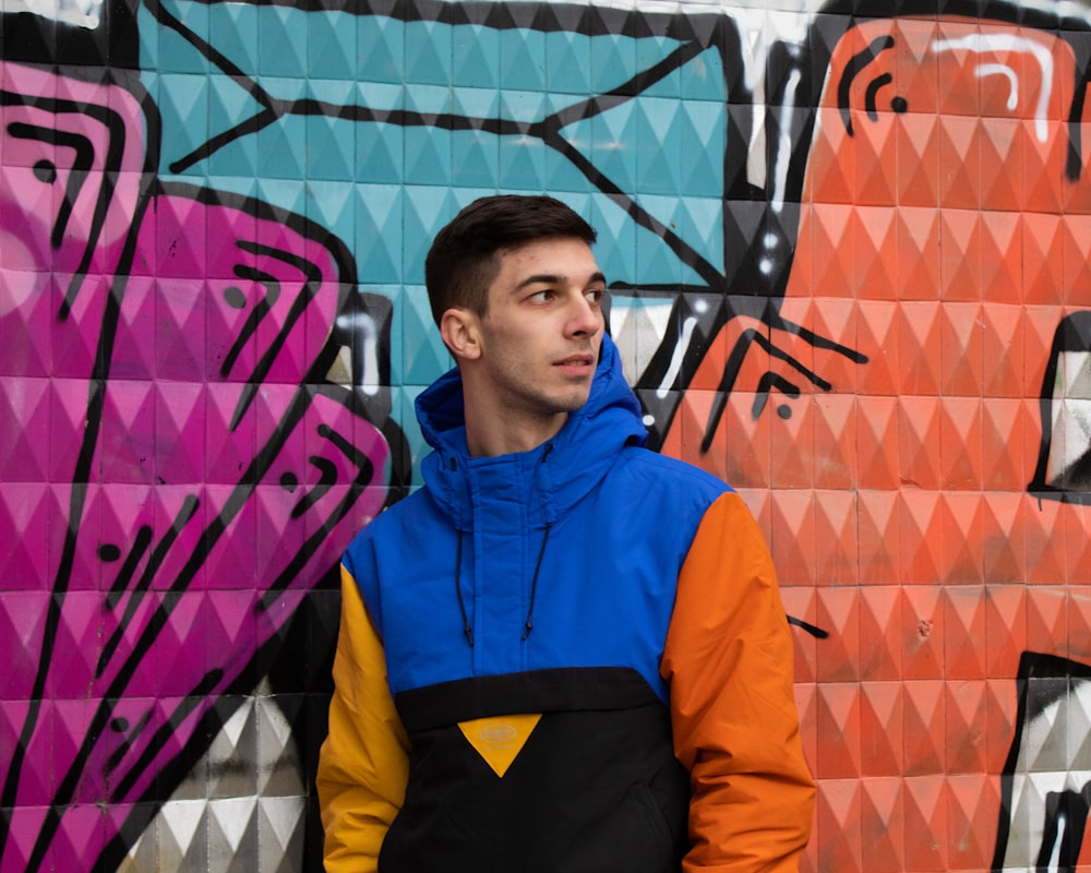 man in orange and blue zip up jacket standing beside wall