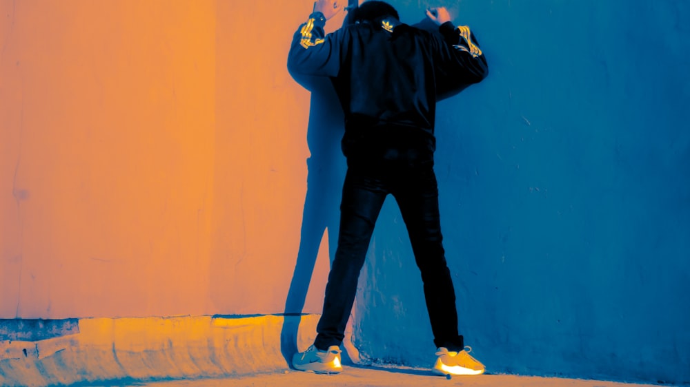 man in black jacket and blue denim jeans standing beside blue wall