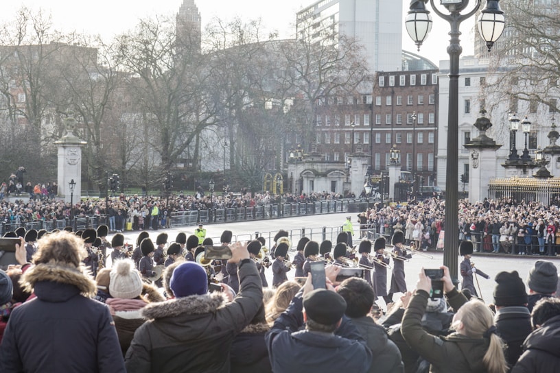 A large crowd gathers on both sides of a street to watch a parade of uniformed soldiers with black hats. The background features historical architecture and bare trees, suggesting it is a cold day. Many spectators capture the event with their phones, indicating a significant public event.