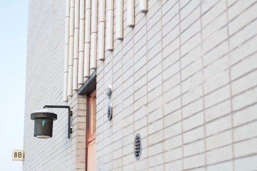 A modern building wall made of light-colored bricks is adorned with a black, cylindrical wall-mounted lamp. The wall features vertical pipe structures and multiple ventilation openings. A door partially visible in the image adds architectural interest. The corner of a number sign, reading '8B', is also seen.