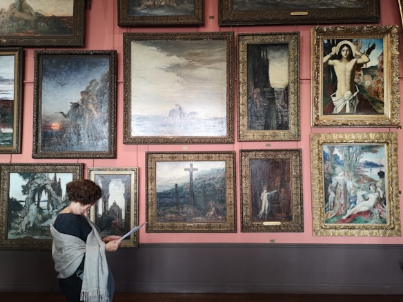 A person stands in front of a gallery wall filled with framed paintings of various sizes. The artworks depict a variety of scenes, including landscapes, a crucifixion, and mythological settings. The wall is painted pink, and the space has a classic, museum-like feel.