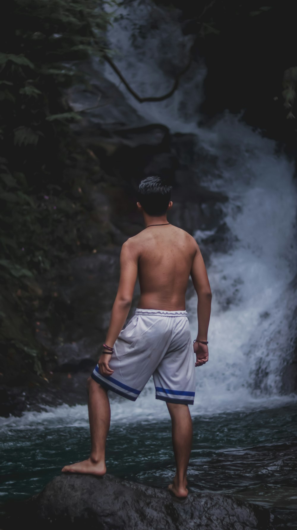 topless man in blue shorts standing on water falls
