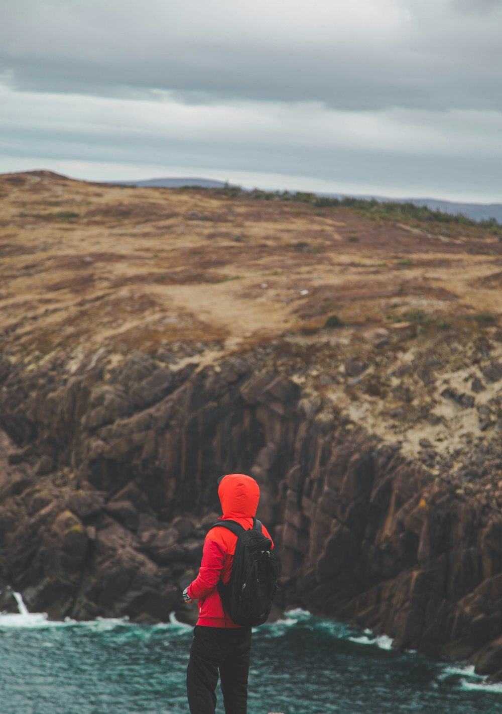 Persona con chaqueta roja y pantalones negros con mochila roja de pie en la montaña rocosa durante el día
