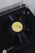 A vinyl record player with a rotating black vinyl disc featuring a yellow label. The label reads 'COEUR DE PIRATE FACE A'. The turntable arm is positioned over the record, and the player itself appears to be sleek and modern in design.