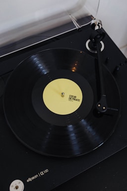 A vinyl record player with a rotating black vinyl disc featuring a yellow label. The label reads 'COEUR DE PIRATE FACE A'. The turntable arm is positioned over the record, and the player itself appears to be sleek and modern in design.