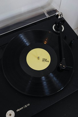 A vinyl record player with a rotating black vinyl disc featuring a yellow label. The label reads 'COEUR DE PIRATE FACE A'. The turntable arm is positioned over the record, and the player itself appears to be sleek and modern in design.