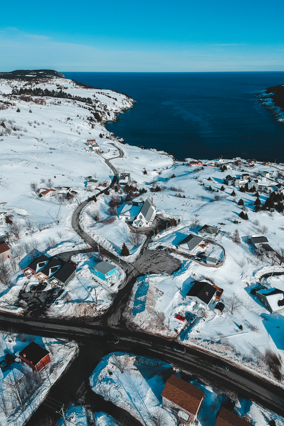 The image presents an aerial view of a small town nestled on a hillside, with houses and buildings scattered across the landscape. The town is surrounded by water, with a body of blue water visible in the background. The sky above is clear and blue, suggesting a bright and sunny day. The snow-covered ground adds to the picturesque winter scene, creating a serene and tranquil atmosphere.