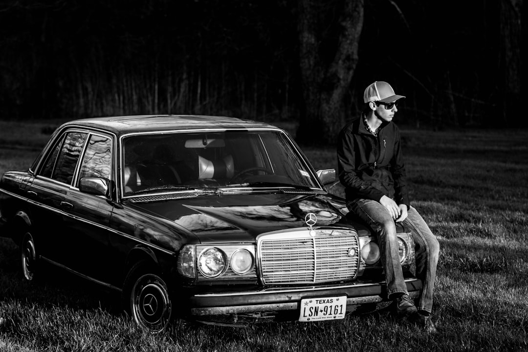 grayscale photo of man in jacket and hat sitting on car