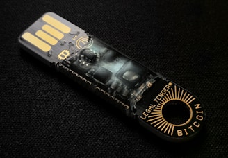 A USB-like device with intricate circuitry and electronic components visible through its transparent casing. The words 'LEGAL TENDER' and 'BITCOIN' are printed near the circular end, featuring gold and black colors.