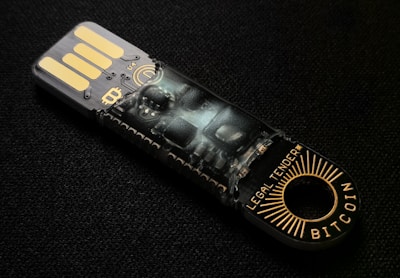A USB-like device with intricate circuitry and electronic components visible through its transparent casing. The words 'LEGAL TENDER' and 'BITCOIN' are printed near the circular end, featuring gold and black colors.