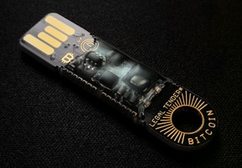 A USB-like device with intricate circuitry and electronic components visible through its transparent casing. The words 'LEGAL TENDER' and 'BITCOIN' are printed near the circular end, featuring gold and black colors.